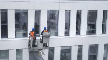Güvenlik kemeri takan iki işçi, duvar, paneller ve ofis binasının ön cephesi önünde vinç beşiği ya da hava platformunda basınçlı temizleyici, silecek ve paspas kullanarak duruyor. Yakın çekim