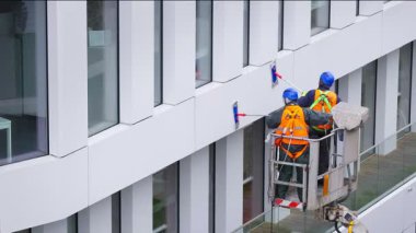 Güvenlik kemeri takan iki işçi, duvar, paneller ve ofis binasının ön cephesi önünde vinç beşiği ya da hava platformunda basınçlı temizleyici, silecek ve paspas kullanarak duruyor. Yakın çekim