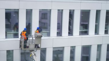 Güvenlik kemeri takan iki işçi, duvar, paneller ve ofis binasının ön cephesi önünde vinç beşiği ya da hava platformunda basınçlı temizleyici, silecek ve paspas kullanarak duruyor. Ön çekim