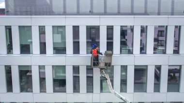 Güvenlik kemeri takan iki işçi, duvar, paneller ve ofis binasının ön cephesi önünde vinç beşiği ya da hava platformunda basınçlı temizleyici, silecek ve paspas kullanarak duruyor. Geniş açı