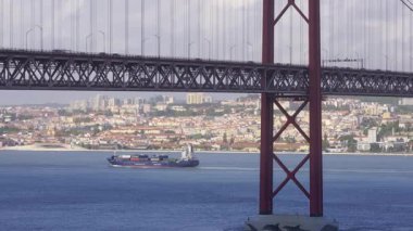 Portekiz, Lizbon 'daki ikonik 25 Nisan köprüsü altında seyreden kargo gemisine yakından bakın. Şehir manzarası, yemyeşil yemyeşil, Tagus nehrinde güneşli bir günde bulutlu gökyüzü. Meşgul deniz ticareti, konteyner gemileri