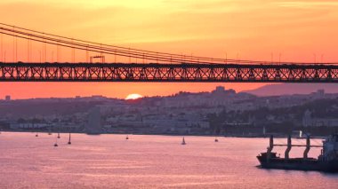 Portekiz, Lizbon 'daki ikonik 25 Nisan köprüsü altında seyreden kargo gemisinin günbatımı görüntüsü. Şehir manzarası, yemyeşil yemyeşil, Tagus nehrinde güneşli bir günde bulutlu gökyüzü. Meşgul deniz ticareti, konteyner gemileri