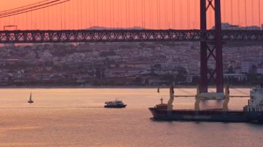 Portekiz, Lizbon 'daki ikonik 25 Nisan köprüsü altında seyreden kargo gemisinin günbatımı görüntüsü. Şehir manzarası, yemyeşil yemyeşil, Tagus nehrinde güneşli bir günde bulutlu gökyüzü. Meşgul deniz ticareti, konteyner gemileri