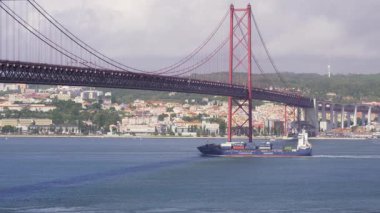 Portekiz, Lizbon 'daki ikonik 25 Nisan köprüsü altında seyreden kargo gemisinin manzarası. Şehir manzarası, yemyeşil yemyeşil, Tagus nehrinde güneşli bir günde bulutlu gökyüzü. Meşgul deniz ticareti, konteyner gemileri
