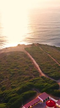 Günbatımında Cabo Espichel veya Cape Espichel 'deki deniz fenerinin insansız hava aracı görüntülerini geri çek. Atlantik Okyanusu kıyısı, Sesimbra, Portekiz. Uç, ufuktan, güneşten, parlaklıktan, yüksek kayalıklardan, Portekizce