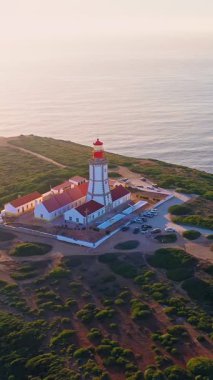 Gün batımında Cabo Espichel veya Cape Espichel 'deki deniz fenerinin hava aracı görüntüsü. Atlantik Okyanusu kıyısı, Sesimbra, Portekiz. Parallaks, etrafta uçmak, yüksek uçurumlar, Portekiz mimarisi, günbatımı yaz manzarası, deniz, güneş ışığı..