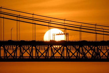 Lizbon, Portekiz 'deki 25 de Abril köprüsünün hemen arkasında batan güneşin yakın görüntüsü, bazı bulutlar, arabalar ve kablolar ile sıcak turuncu gökyüzü parlayan güneşe karşı, çarpıcı kentsel günbatımı sahnesi