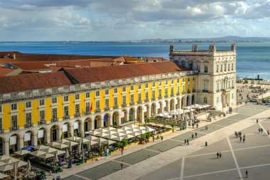 Portekiz, Lizbon 'daki Ticaret Meydanı veya Praca do Comercio' nun manzarası, ikonik sarı Pombaline mimarisi, hareketli açık hava kafeleri, Tagus Nehri, şehir yaşamı, tarihi şehir merkezi, güneşli gün, gökyüzü