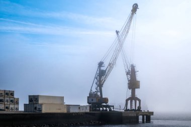 Rıhtımda büyük endüstriyel vinçler sabah sisli konteynırların yanında. Sisli rıhtım sahnesi, güneşli bir günde şafak vakti yoğun kargo limanının deniz sanayisi, lojistik ve atmosferini vurguluyor
