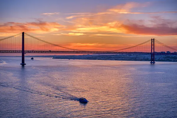 Portekiz, Lizbon 'da Tagus Nehri üzerinde siluet halinde 25 de Abril Köprüsü üzerinde gün batımında sakin sularda yüzen küçük bir tekneyle. Canlı turuncu ve mor gökyüzü, dramatik arkaplan, ünlü Portekiz simgesi, renkli yansımalar, manzara, yaz sisi 