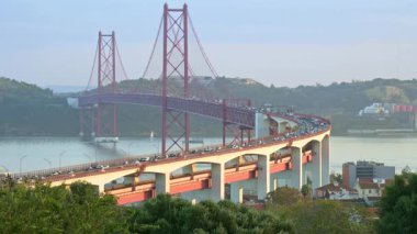 Tagus Nehri üzerindeki 25 de Abril Köprüsü 'nün bulutlu bir günde gün batımında yoğun trafik varken Lizbon' un ikonik manzarası. Şehir trafiği, yemyeşil yeşillik, sakin nehir, ünlü simgeler, kırmızı asma köprü, renkli gökyüzü, seyahat yeri.