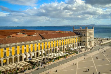 Portekiz, Lizbon 'daki Commerce Square veya Praca do Comercio' nun geniş açılı görüntüleri, ikonik sarı Pombaline mimarisi, hareketli açık kafeler, Tagus Nehri, şehir yaşamı, tarihi şehir merkezi, güneşli bir gün, bulutlu gökyüzü. Yaz tatili, tatil beldesi