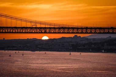 25 de Abril köprüsünün üzerinde silüet ve Tagus nehrinde gün batımında aşağıda sürüklenen yelkenliler ve arkada yaz sisi içinde Lizbon silueti. Turuncu gökyüzü, bulutlar ve güneş, sakin sular, köprüdeki trafik, Lizbon, Portekiz 'in ikonik manzarası.