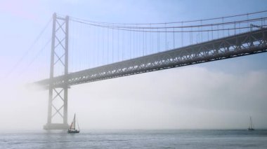 25 Nisan 'da, Tagus Nehri, Lizbon, Portekiz üzerindeki yoğun siste güneşli bir günde geçen yelkenli teknelerinin statik görüntüleri. Üzerinde sis olan güzel asma köprü manzarası, sessiz tonlar, yumuşak aydınlatma, sakin nehir suları, berrak mavi gökyüzü..