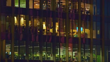 Geceleri modern ofis binası. Büyük camlı pencereleri var. Çalışma alanlarını sıcak aydınlatıyor. Çalışanlar masalarda ve bilgisayarlarda, aktif iş ortamında ve günbatımından sonra modern iş ortamında görülebilirler.