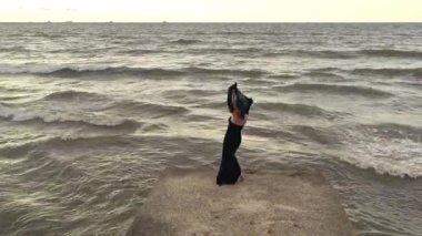 İskelede dans eden atkısı olan zarif bir kadın. İHA görüntüsü. Dalgalı deniz. Siyah uzun elbiseli çekici bayan..