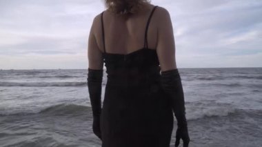 İskelede dans eden atkısı olan zarif bir kadın. İHA görüntüsü. Dalgalı deniz. Siyah uzun elbiseli çekici bayan..