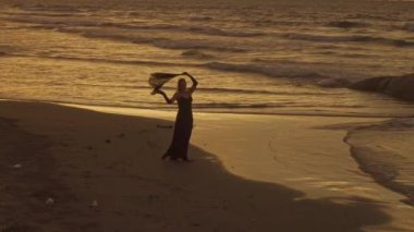 İskelede dans eden atkısı olan zarif bir kadın. İHA görüntüsü. Dalgalı deniz. Siyah uzun elbiseli çekici bayan..