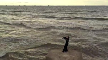 İskelede dans eden atkısı olan zarif bir kadın. İHA görüntüsü. Dalgalı deniz. Siyah uzun elbiseli çekici bayan..