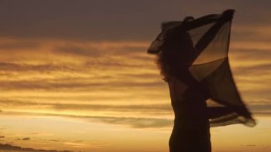 Genç mutlu romantik kız günbatımı kumsalında ayakta duruyor ya da dans ediyor. Kollarını kaldırıp eşarp takıyor.