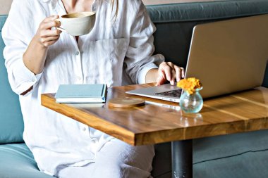 Elinde bir fincan kahve olan bir kadın laptopun başında tanınmadan çalışıyor.