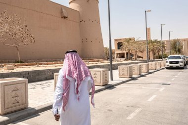 Arap adam, geleneksel Arap kıyafetleri giyip Riyad Suudi Arabistan 'daki El Masmak Sarayı Müzesi' nin yanında geziyor.