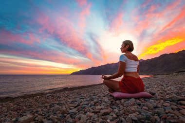 Genç kadın Shilhouette, gün batımına karşı sahilde meditasyon pozisyonunda.