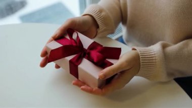 Kadın elleri hediyeyi tutuyor. Yumuşak pembe renkleri kapatıyor. Kırmızı kurdele.