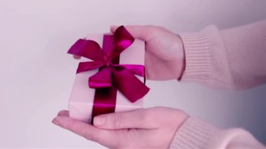 Kadın elleri hediyeyi tutuyor. Yumuşak pembe renkleri kapatıyor. Kırmızı kurdele.