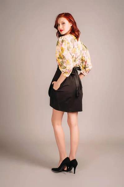 Female fashion. Redhead lady in elegant mini evening dress with long yellow balloon sleeves and black skirt on grey studio background. Young woman with red lips and natural hairdo. Holiday outfit.