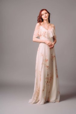 Çiçek desenli bej bir gece elbisesi giyen çekici genç bir kadın. Gri arka planda gülümseyen kızıl bayanın stüdyo portresi. Omuzlar büyük önlükten, zarif bir görünüş. Kadın yaz kıyafeti..