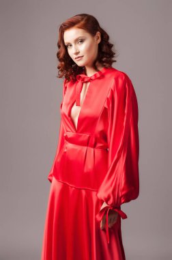 Kırmızı yakalı uzun kollu saten maksi elbise. Büyük beden şenlik elbisesi. Gri stüdyo arka planında gülümseyen parlak kızıl kadının portresi. Zarif modern bohem kadın görünüşü