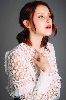 Süslü, beyaz, ilham verici kadın kıyafeti içinde uzun kollu bir bluz. Gri arka planda güzel, kızıl saçlı bir kadının stüdyo portresi. Kırmızı dudaklı doğal makyaj ve saç modeli..