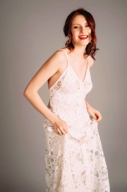 Gülen kızıl saçlı kadının gizli stüdyo resmi. Nakışlı ve derin dekolteli beyaz gelinlik. Kolsuz, ince bir gece elbisesi, gelinlik. Doğal makyaj ve kıvırcık saç, kırmızı dudaklar.