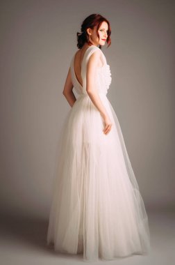 Muhteşem mutlu genç gelinin stüdyo portresi. Çiçeklerle süslenmiş beyaz tül elbise. V yakalı gelinlik, kesilmiş. Klasik bir gelinlik. Kızıl saçlı, kolsuz maksimum balo elbisesi giyen, gülümseyen bir kadın.