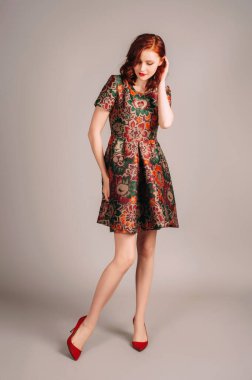 Kahverengi gece elbisesi ve kırmızı topuklularla yürüyen güzel kızıl saçlı genç kadının stüdyo portresi. Zarif moda parlak sonbahar renkleriyle görünür. Jacquard çiçekli özel dikim mini elbise.