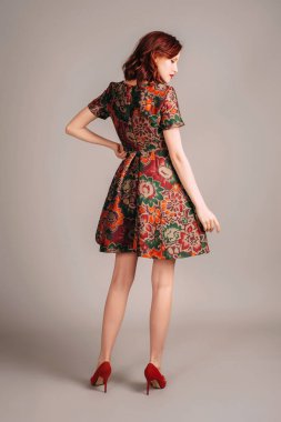 Kahverengi gece elbisesi ve kırmızı topuklularla yürüyen güzel kızıl saçlı genç kadının stüdyo portresi. Zarif moda parlak sonbahar renkleriyle görünür. Jacquard çiçekli özel dikim mini elbise.