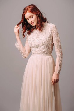 Fildişi uzun kollu Maxi gelinlik. Çiçekli nakış ve tül etekli şeffaf korse. Gülümseyen kızıl saçlı gelinin şehvetli portresi. Moda bir gelin saç stili ve makyaj. Nedimenin görünüşü