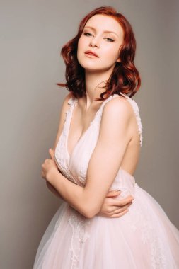 Kolsuz, rengarenk, pembe bir balo elbisesi, koyu seksi yaka, sabitlenmiş kayışlar. Sevimli kızıl saçlı gelinin şehvetli stüdyo portresi. Şık tül gelinlik, modaya uygun dalgalı saç stili.