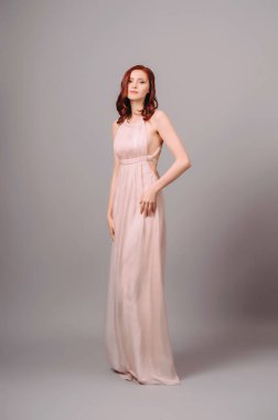 Pastel transformatör elbise. Baştan çıkarıcı kızıl saçlı kadın, maksimum gece elbisesi ve gri stüdyo arka planında yüksek topuklu ayakkabılar. Nedimenin yaz ipek elbisesi. Kıvırcık saçlı, güzel, seksi bir bayan.