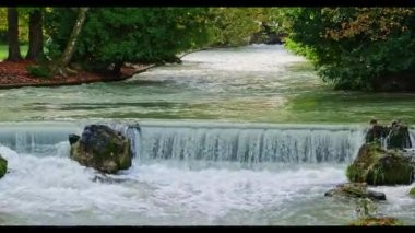 İngiliz Bahçesi, Münih, Almanya 'daki Eisbach nehrinde küçük bir şelale yeşil yosun kayalarının üzerinden dökülen ve uzun yeşil ağaçlarla çevrili isar dalgaları.