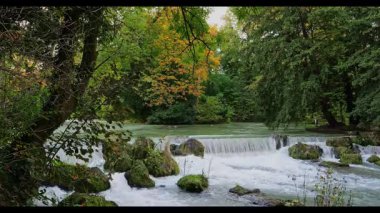 İngiliz Bahçesi, Münih, Almanya 'daki Eisbach nehrinde küçük bir şelale yeşil yosun kayalarının üzerinden dökülen ve uzun yeşil ağaçlarla çevrili isar dalgaları.
