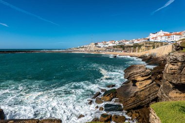Portekiz 'in Ericeira kentindeki Praia do Sul manzarası altın kum, turkuaz sular ve canlı bir gökyüzü altında kıyı binaları.