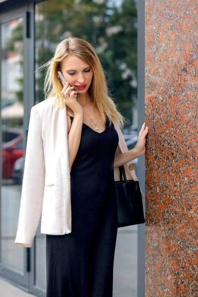 A young attractive blonde girl in a black dress and an oatmeal jacket communicates on an iPhone. Background - urban modern architecture in summer. Communication and women concept