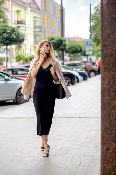 A tall blonde in a black dress and an oatmeal-colored jacket on the shoulders is talking on a mobile phone while walking down the street. Background of the city during the day. Modern life concept