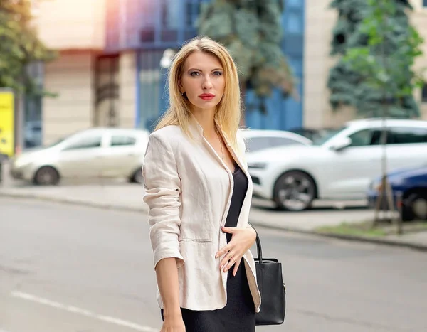 Women. Business concept. Portrait with an attractive blonde business lady in an oatmeal jacket and a black dress with an iPhone in her hand on the street. Summer urban landscape - Background.