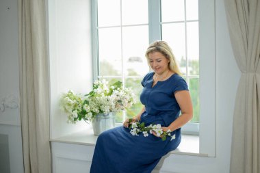 Mavi elbiseli 40 yaş üstü güzel bir kadın pencerenin kenarındaki pencere pervazında çiçeklerle oturuyor. Bahar konsepti