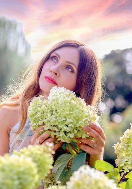 A stunning portrait of a beautiful Caucasian girl in a light flower print dress in a garden where flowers bloom. Summer. Beauty concept.	