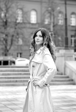 Bir şehir binasının kırmızı cephesine karşı bej yağmurluk giymiş zarif esmer bir kızın siyah beyaz portresi. Sonbahar. Moda konsepti