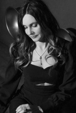 Esmer, beyaz bir kadının siyah ve beyaz portresi, soluna özgüvenli bir bakış, yüzüne yakın bir el, çatı katı tarzı bir iç mekanda siyah bir şezlongda oturuyor. Güzellik ve f